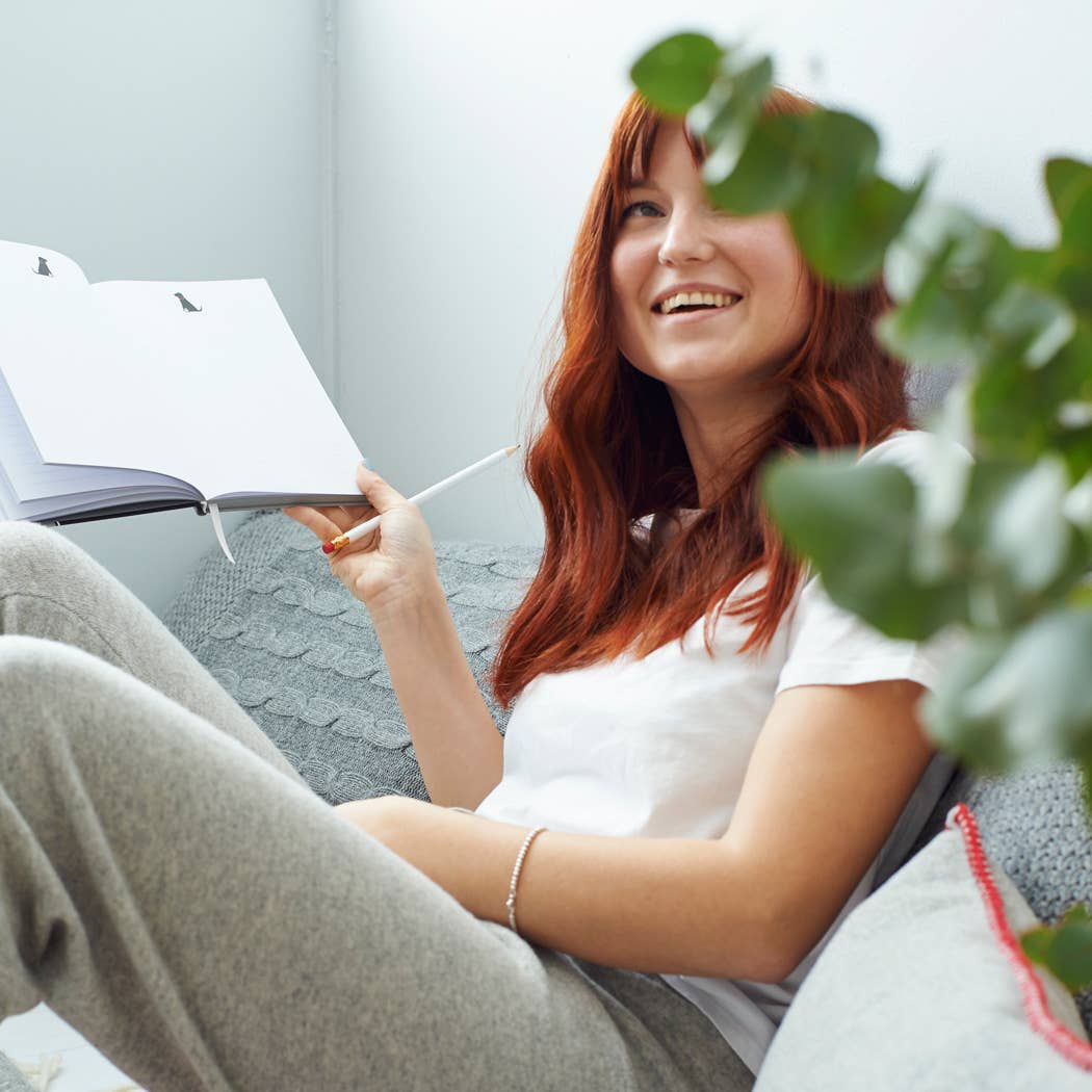 A smiling woman with red hair sits on a gray couch holding an open notebook and white pen, wearing a white t-shirt and gray sweatpants, with a green leafy plant in the foreground.