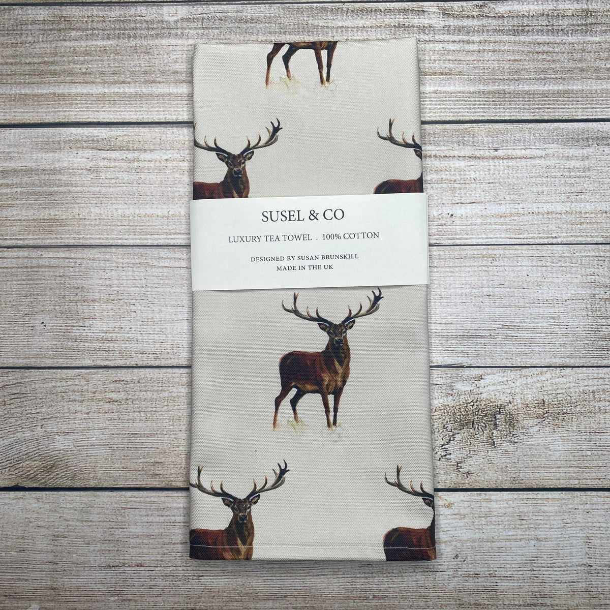 A folded cream-colored cotton tea towel with a printed deer pattern, wrapped in a white paper band labeled 'SUSEL & CO', placed on a rustic gray wooden plank background.
