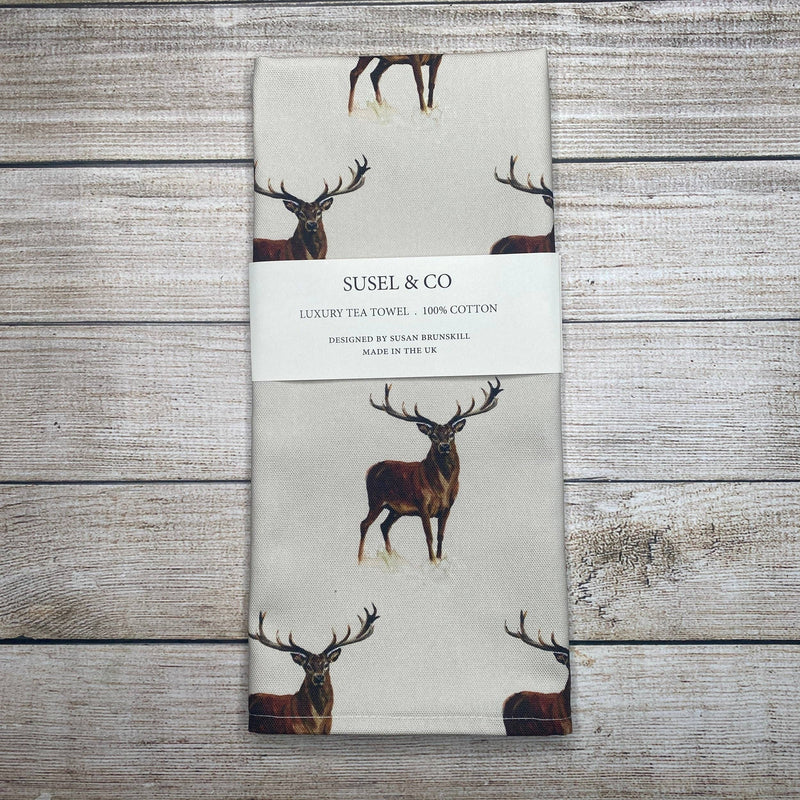 A folded cream-colored cotton tea towel with a printed deer pattern, wrapped in a white paper band labeled 'SUSEL & CO', placed on a rustic gray wooden plank background.
