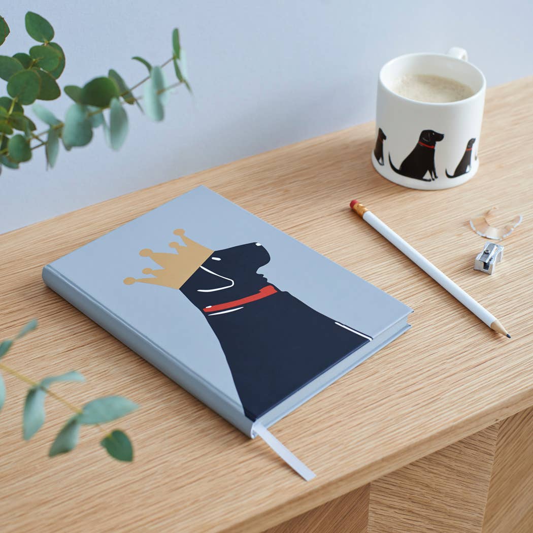 A gray hardcover notebook with a stylized black dog wearing a gold crown and red collar, styled on a wooden desk next to a white coffee mug, pencil, and eraser.