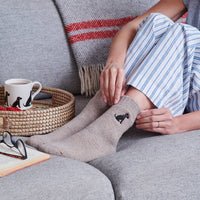 A person sitting on a gray couch wearing taupe socks with a small black cat embroidered on the side. A woven tray with a white mug, a book, and glasses sits beside them, with a striped shirt and blanket visible in the background.