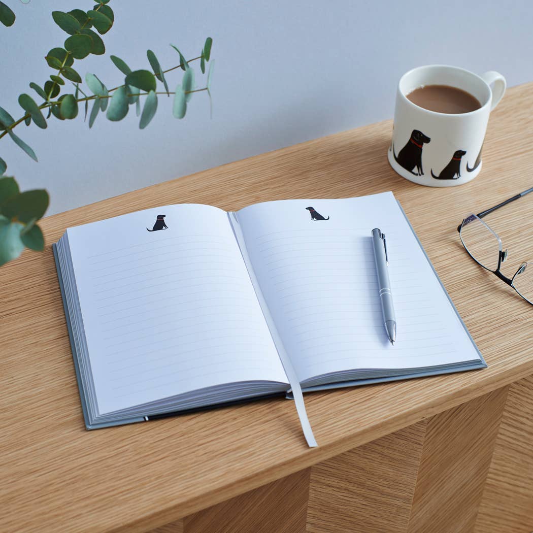 An open lined notebook with small black dog silhouettes on the pages sits on a wooden table, styled with a silver pen, a white coffee mug with black dog illustrations, and eyeglasses. A green plant sprig is visible in the upper left corner.
