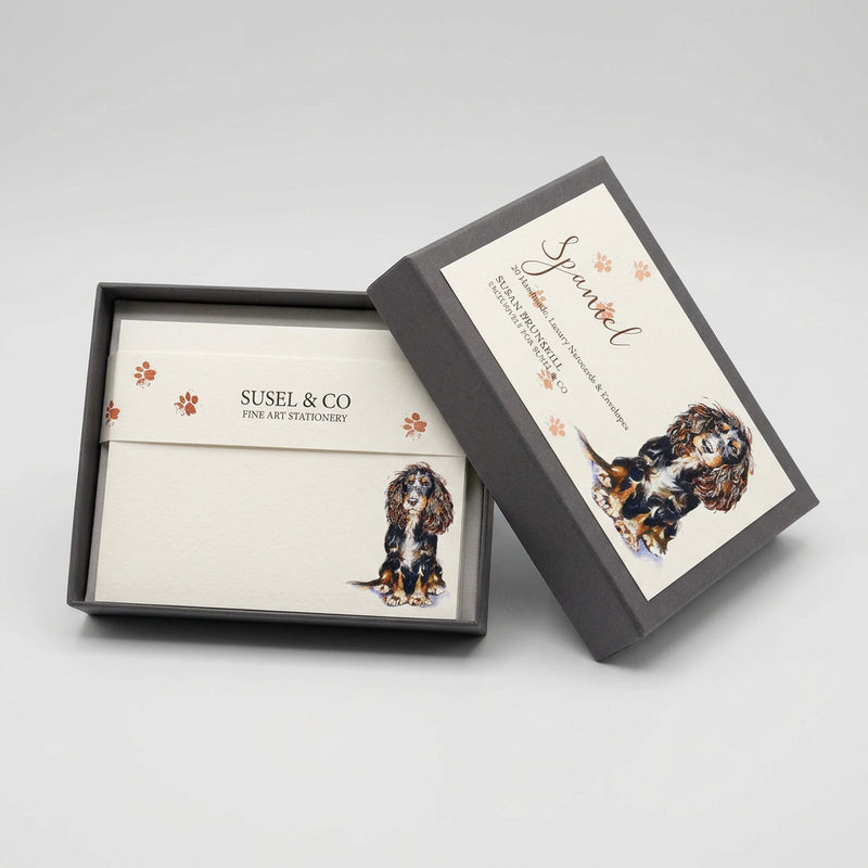 Two framed custom pet memorial cards displayed on a white wall above a gray fabric surface. The cards feature watercolor illustrations of a black and white Cocker Spaniel and are personalized with the name 'Spencer' and the brand 'SUSEL & CO'.
