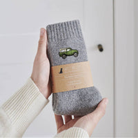 A person holds a pair of gray ribbed socks with a green jeep graphic and a kraft paper band labeled 'SWEET CHILLIONS'. The background is a clean, off-white paneled wall.