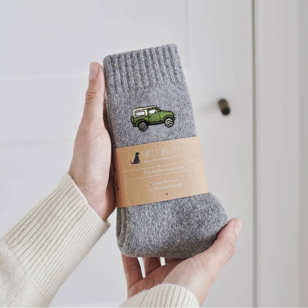 A person holds a pair of gray ribbed socks with a green jeep graphic and a kraft paper band labeled 'SWEET CHILLIONS'. The background is a clean, off-white paneled wall.
