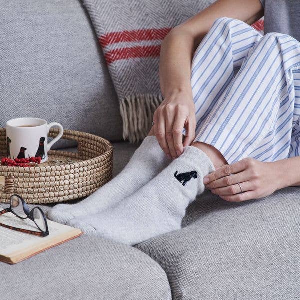 Person wearing light gray socks with a black cat design, sitting on a couch with a mug and glasses nearby.