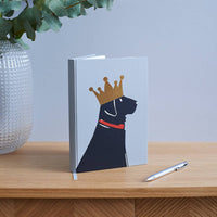 A hardcover notebook with a minimalist illustration of a black dog wearing a gold crown and red collar, standing on a wooden table next to a silver pen with a vase of eucalyptus in the background.