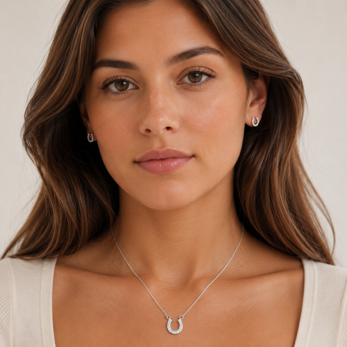 A woman with brown hair wearing a delicate silver horseshoe pendant necklace and matching stud earrings, shown in a close-up portrait against a soft light background.