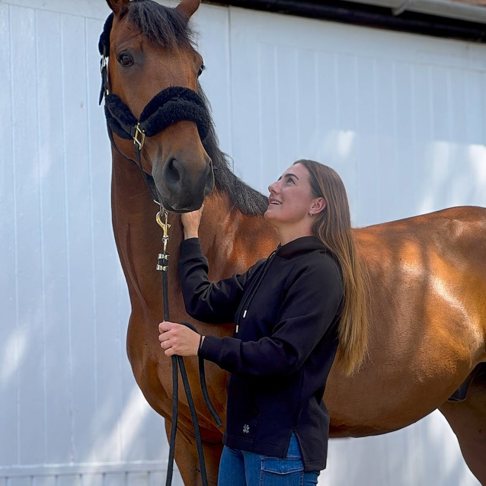 Woman in equestrian attire standing next to a horse in front of a white wall.