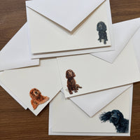 A set of white stationery cards and matching envelopes arranged on a wooden surface, each printed with a different dog portrait illustration in black, brown, and orange-brown tones.