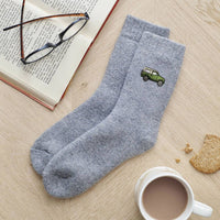 A pair of heather gray crew socks with a small green embroidered car on the side, arranged on a light wood tabletop with an open book, eyeglasses, a cup of coffee, and a cookie as props.