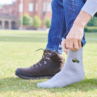A person outdoors on grass wearing a heather gray sock with a small green truck graphic near the ankle, paired with blue jeans and a dark brown lace-up boot.