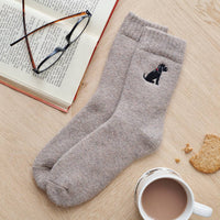 A pair of heather taupe knit crew socks with a black dog embroidered on the cuff, styled on a light wood tabletop with an open book, eyeglasses, a cup of tea, and a biscuit.