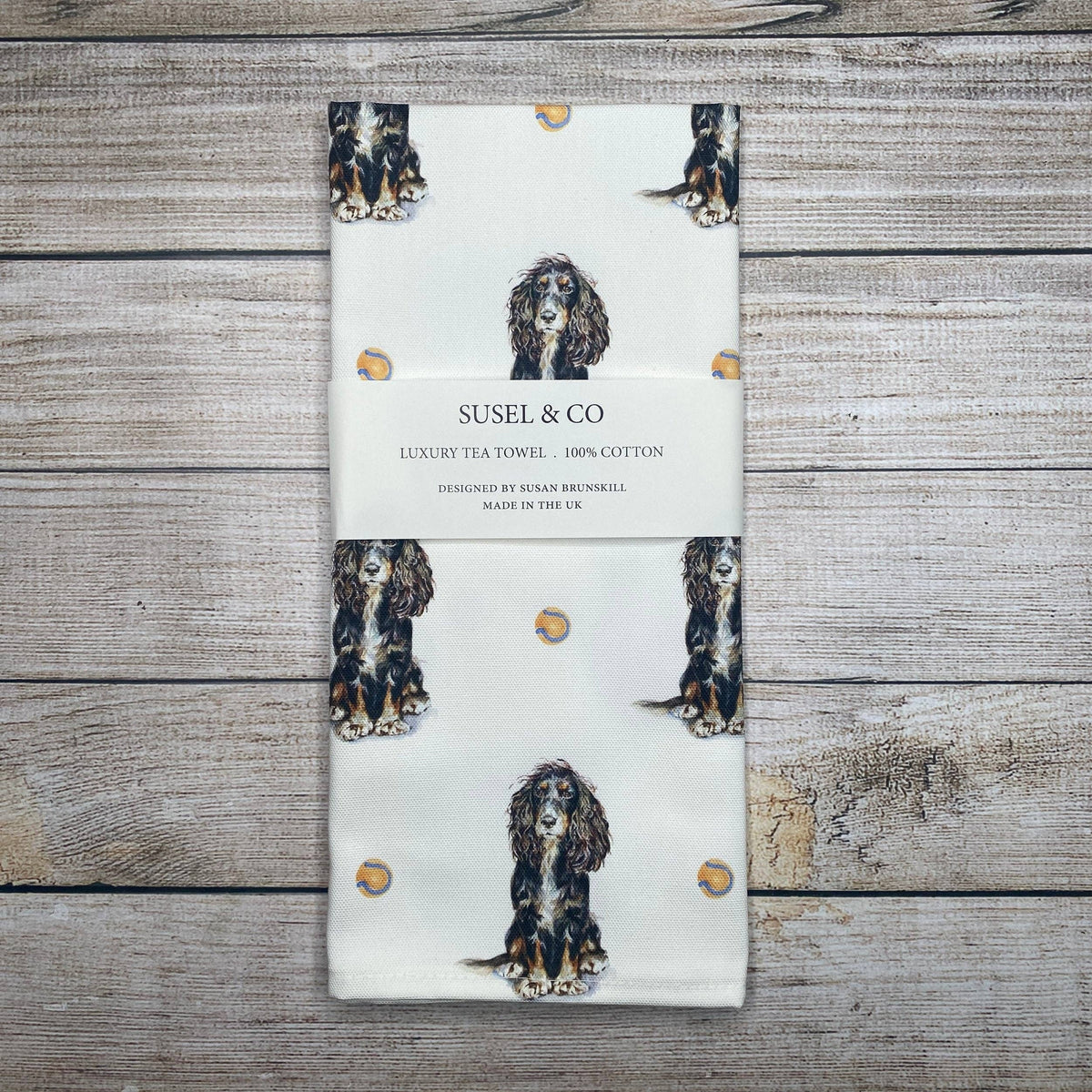 A folded cotton tea towel with a white background, printed with illustrations of three-legged dog portraits and small gold dots, wrapped in a paper band labeled 'SUSEL & CO'. The towel is photographed flat on a rustic, light-colored wooden plank background.