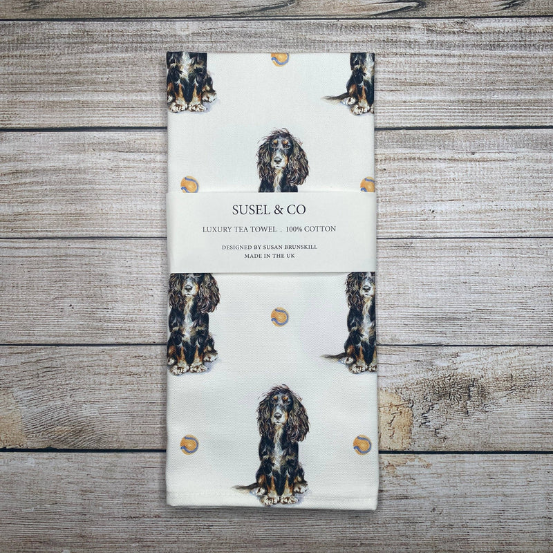 A folded cotton tea towel with a white background, printed with illustrations of three-legged dog portraits and small gold dots, wrapped in a paper band labeled 'SUSEL & CO'. The towel is photographed flat on a rustic, light-colored wooden plank background.