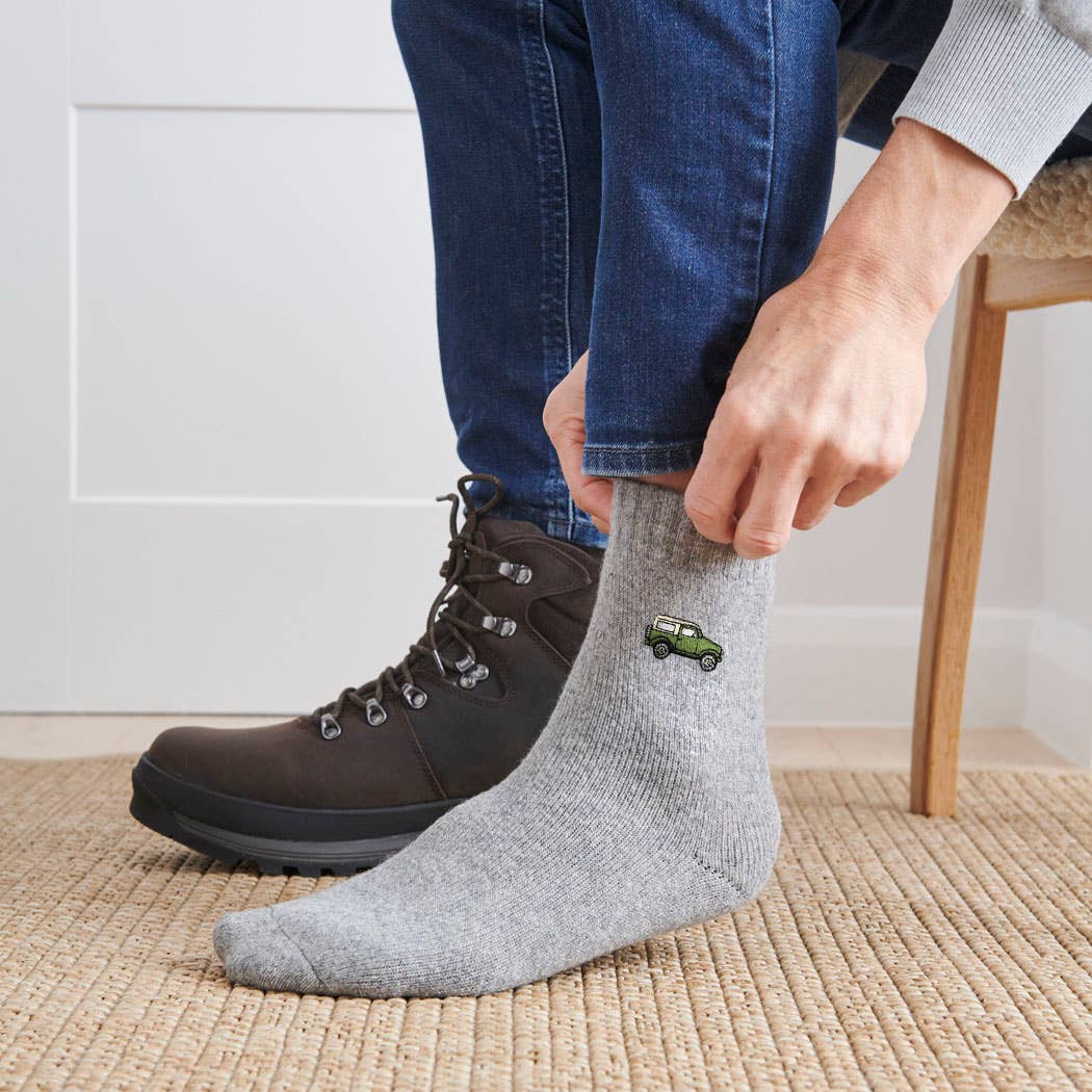 A person wearing blue jeans, a brown lace-up hiking boot, and a light gray crew sock with a small green car graphic, standing on a woven rug indoors.
