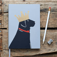 A hardcover notebook with a light blue cover featuring a black dog wearing a gold crown and red collar, styled on a rustic wooden surface next to a white pencil and a metal pencil sharpener.