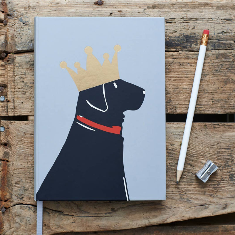 A hardcover notebook with a light blue cover featuring a black dog wearing a gold crown and red collar, styled on a rustic wooden surface next to a white pencil and a metal pencil sharpener.