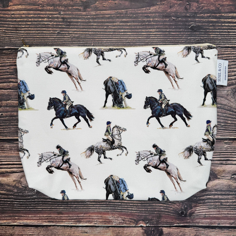 A zippered pouch with an off-white background and a repeating print of equestrian illustrations, laid flat on a dark wood surface. A small fabric tag on the right reads 'EQUESTRIAN & CO'.