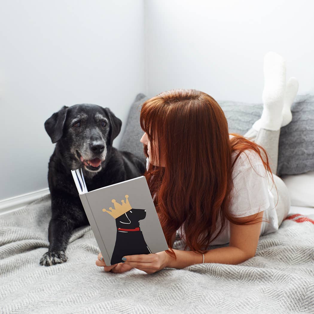 A woman with long red hair lies on a bed reading a book with a black dog wearing a gold crown illustration on the cover, while a real black dog sits beside her.